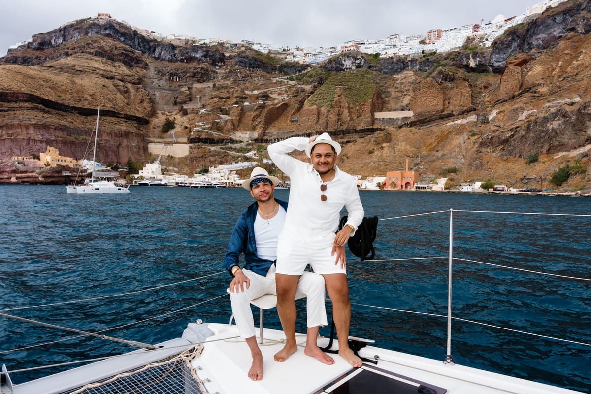 Two men on a catamaran bow with the old port and cliffs of Fira behind, Santorini