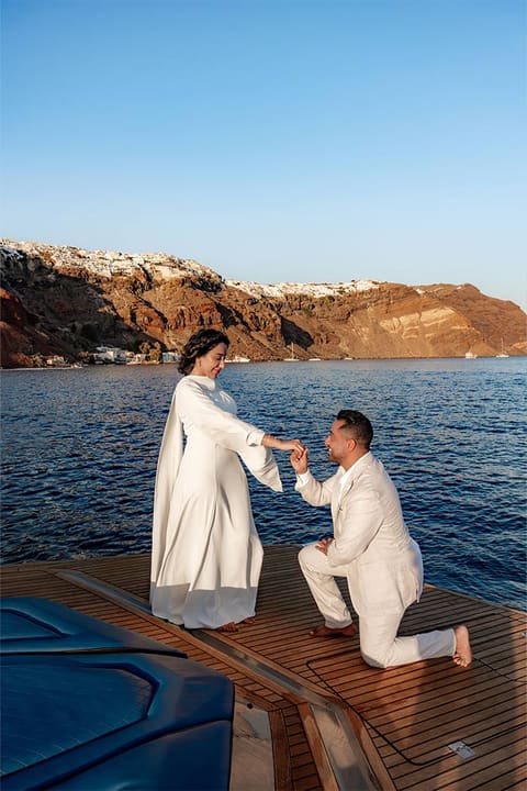 Man on one knee on a catamaran deck below the cliffs of Oia, proposing in Santorini