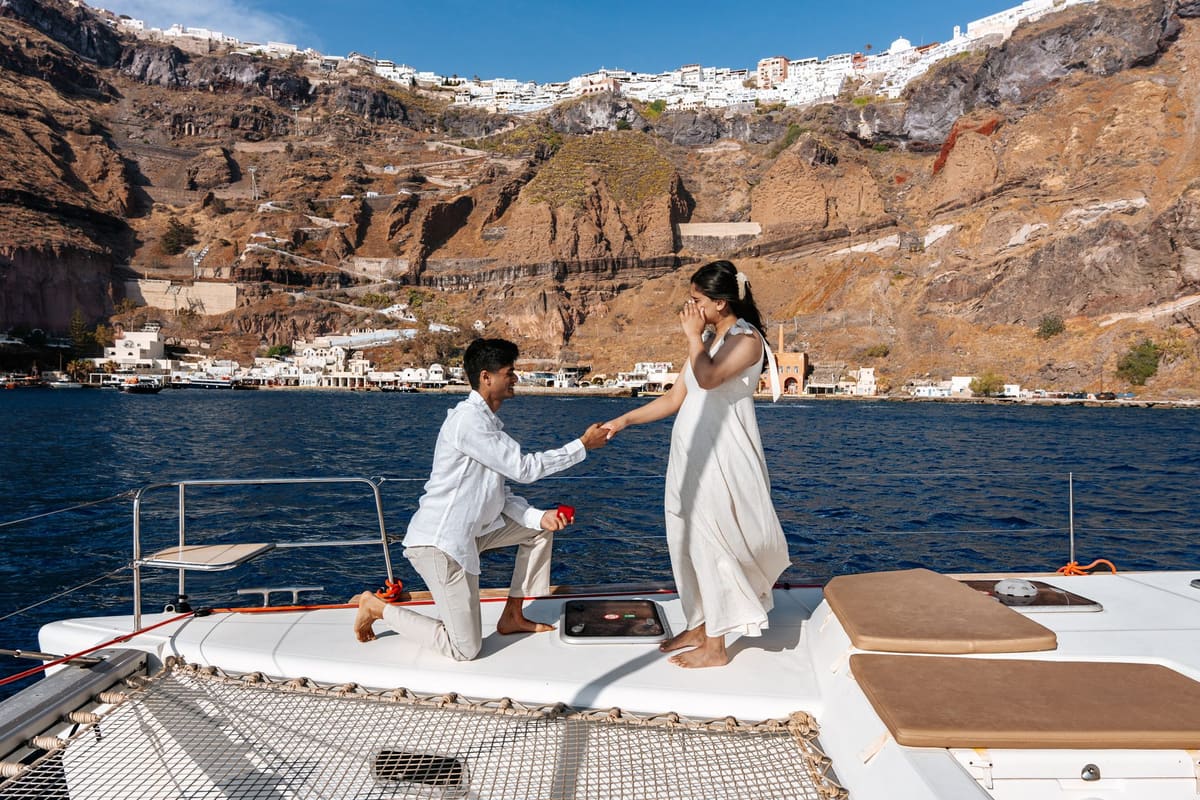 Man kneeling on the catamaran bow to propose below the cliffs of Oia, Santorini