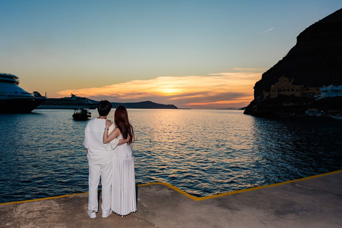 Couple on the pier of the old port of Fira at sunset, the caldera cliffs behind
