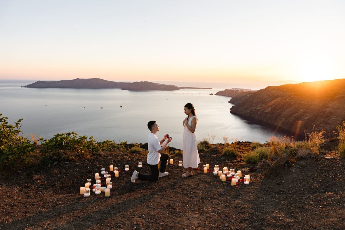 Man on one knee among candles on a cliff path above the Santorini caldera at sunset